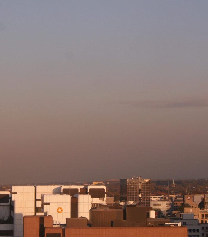L'horizon d'une ville au coucher du soleil, avec des immeubles de bureaux modernes à gauche et des clochers historiques à droite, le tout sous un ciel clair avec une lueur chaude et dorée.