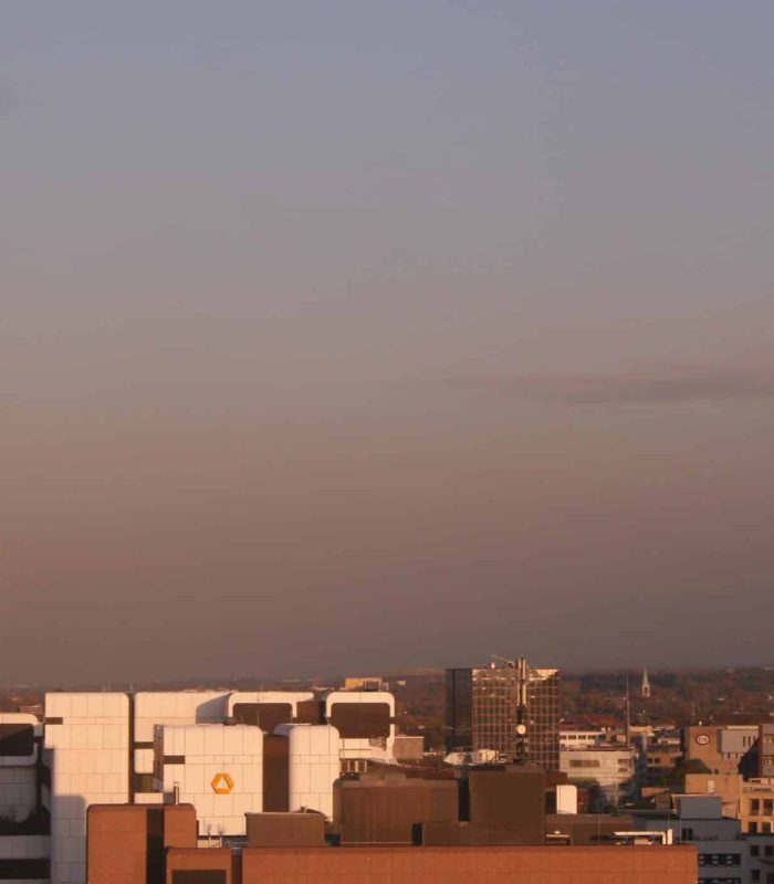 A city skyline at sunset, with modern office buildings on the left and historic bell towers on the right, all under a clear sky with a warm, golden glow.