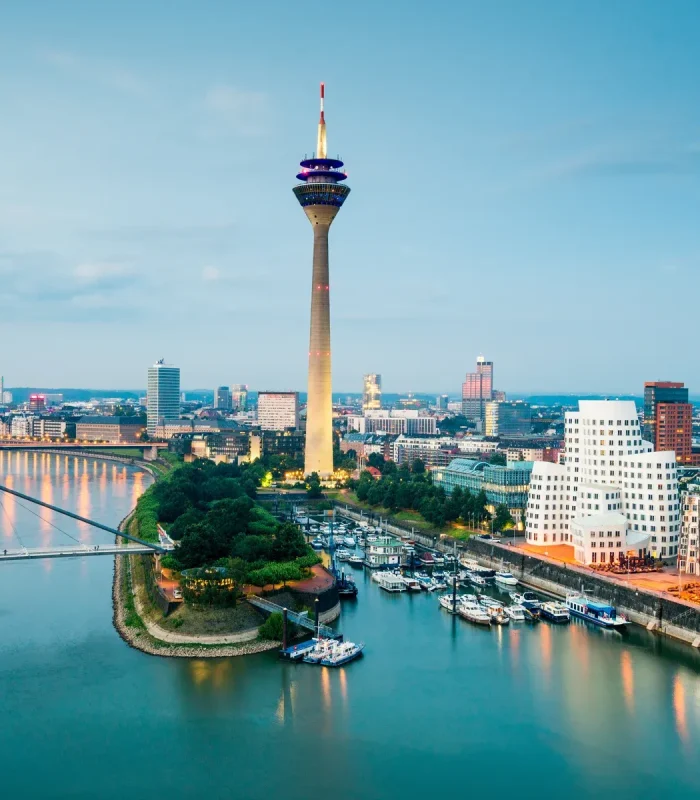 Vue de la ville de Düsseldorf, en Allemagne, avec le Rhin, la tour de télévision Rheinturm, des bâtiments modernes et des bateaux amarrés au bord de l'eau au crépuscule.