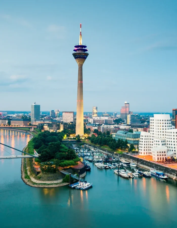 Vue de la ville de Düsseldorf, en Allemagne, avec le Rhin, la tour de télévision Rheinturm, des bâtiments modernes et des bateaux amarrés au bord de l'eau au crépuscule.