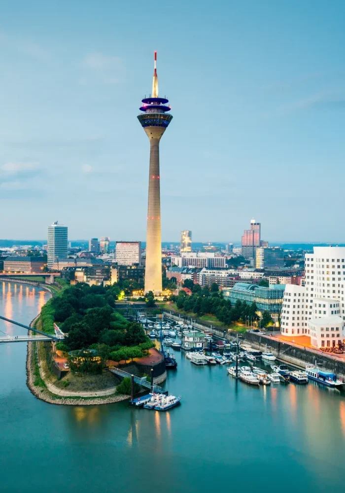 Vue de la ville de Düsseldorf, en Allemagne, avec le Rhin, la tour de télévision Rheinturm, des bâtiments modernes et des bateaux amarrés au bord de l'eau au crépuscule.