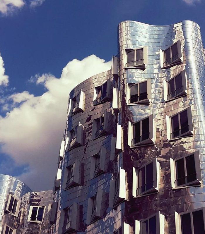 Un bâtiment moderne avec une façade ondulée en métal argenté réfléchissant et de nombreuses fenêtres, sur fond de ciel bleu avec des nuages blancs épars.