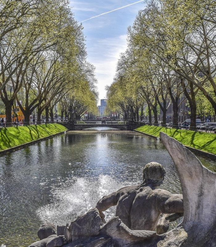 Un canal bordé d'herbe verte et d'arbres des deux côtés, avec une statue en pierre et une fontaine au premier plan. Des voitures sont garées le long de la route et des bâtiments sont visibles à l'arrière-plan sous un ciel dégagé.