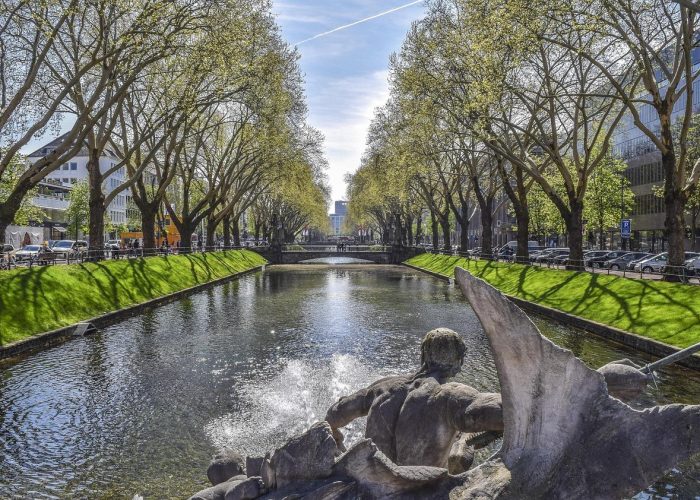 Un canal bordé d'herbe verte et d'arbres des deux côtés, avec une statue en pierre et une fontaine au premier plan. Des voitures sont garées le long de la route et des bâtiments sont visibles à l'arrière-plan sous un ciel dégagé.