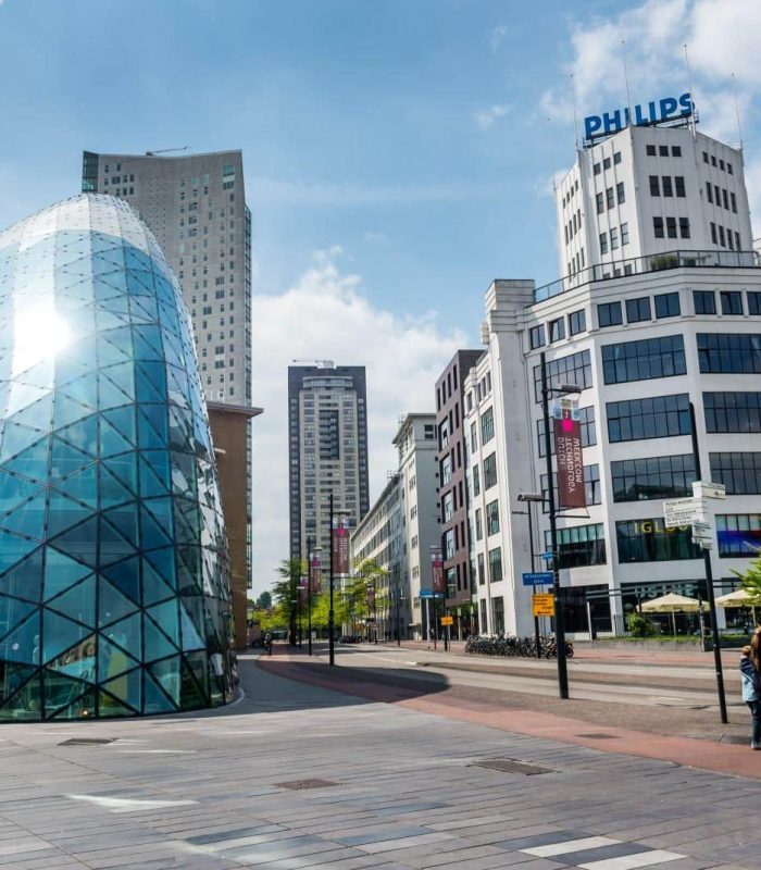 Place d'une ville moderne avec, à gauche, un bâtiment en verre en forme de dôme et, à droite, un grand bâtiment blanc portant l'enseigne PHILIPS ; des piétons marchent dans une large rue sous un ciel partiellement nuageux.
