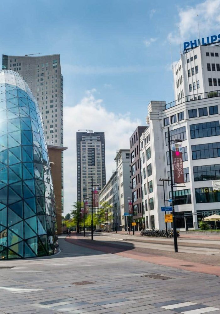 Place d'une ville moderne avec, à gauche, un bâtiment en verre en forme de dôme et, à droite, un grand bâtiment blanc portant l'enseigne PHILIPS ; des piétons marchent dans une large rue sous un ciel partiellement nuageux.