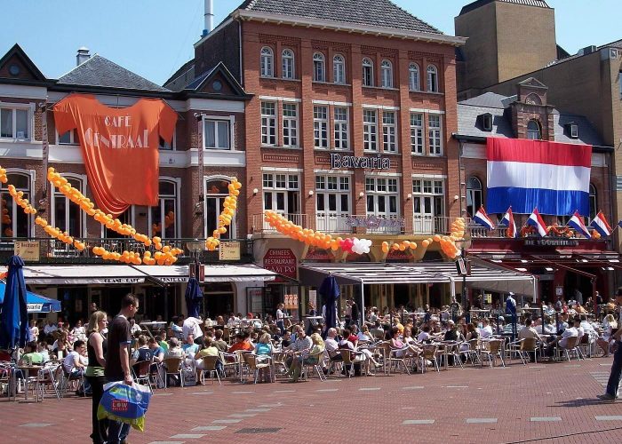 Une place extérieure animée avec de nombreuses personnes assises à des tables de café. Des drapeaux néerlandais et des décorations orange sont accrochés aux bâtiments en briques rouges, suggérant un événement festif aux Pays-Bas.