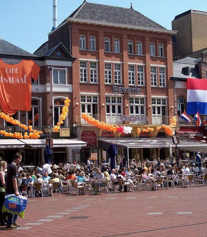 Une place extérieure animée avec de nombreuses personnes assises à des tables de café. Des drapeaux néerlandais et des décorations orange sont accrochés aux bâtiments en briques rouges, suggérant un événement festif aux Pays-Bas.