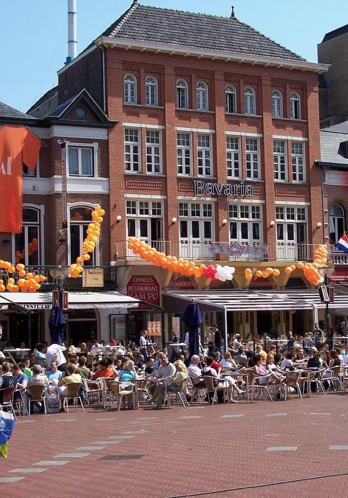 Une place extérieure animée avec de nombreuses personnes assises à des tables de café. Des drapeaux néerlandais et des décorations orange sont accrochés aux bâtiments en briques rouges, suggérant un événement festif aux Pays-Bas.