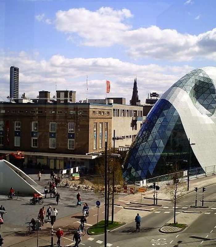Un bâtiment moderne à dôme de verre et au design géométrique se dresse à côté de structures plus anciennes sur une place de ville animée, où les gens se promènent à pied ou à vélo sous un ciel partiellement nuageux.