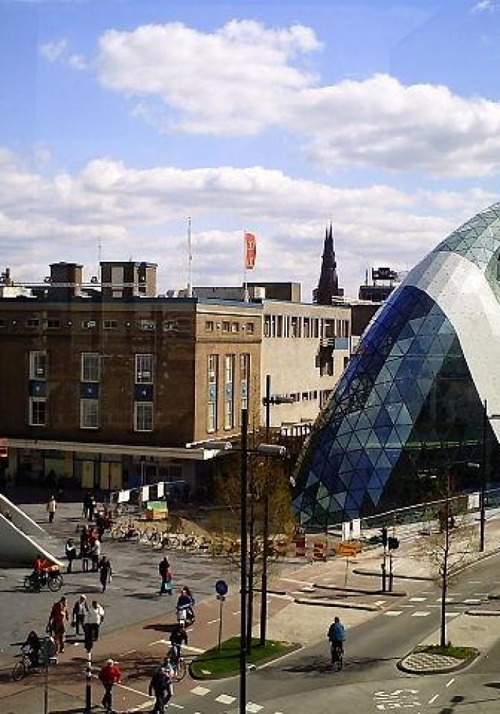 Un bâtiment moderne à dôme de verre et au design géométrique se dresse à côté de structures plus anciennes sur une place de ville animée, où les gens se promènent à pied ou à vélo sous un ciel partiellement nuageux.
