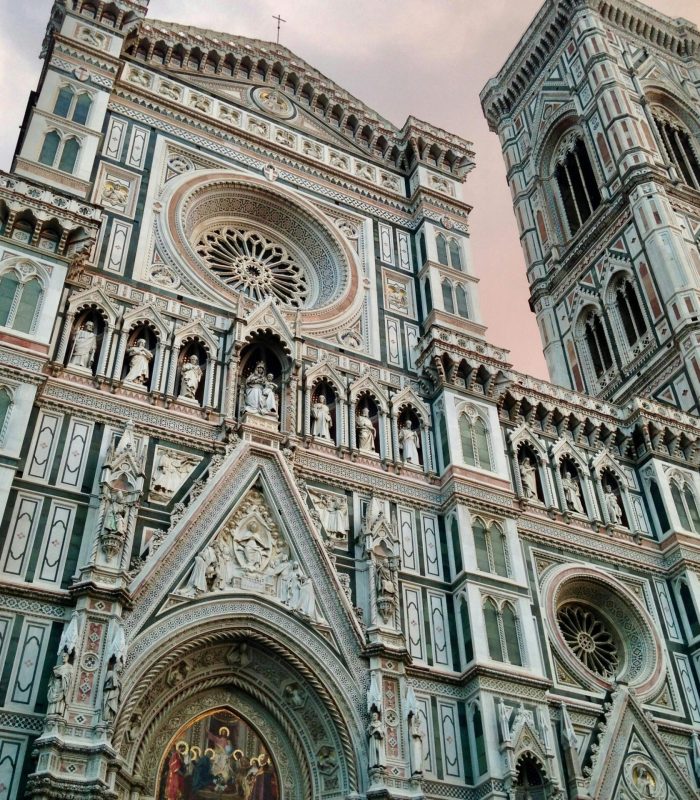 La façade et le clocher en marbre de la cathédrale de Florence, avec ses sculptures détaillées, ses arcs et ses rosaces, sur fond de ciel nuageux.