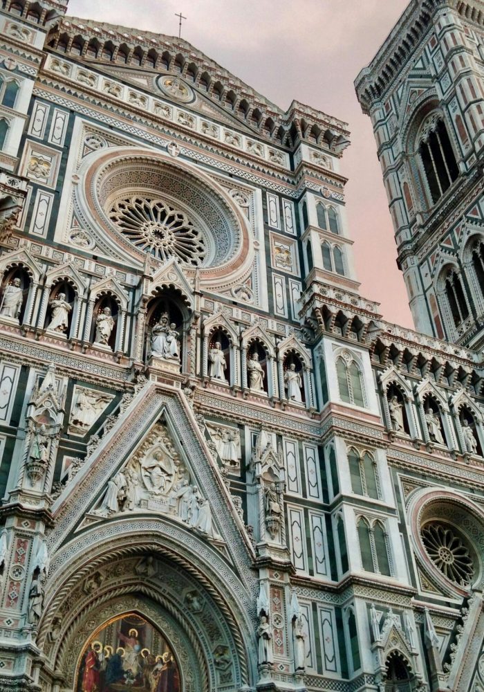 La façade et le clocher en marbre de la cathédrale de Florence, avec ses sculptures détaillées, ses arcs et ses rosaces, sur fond de ciel nuageux.