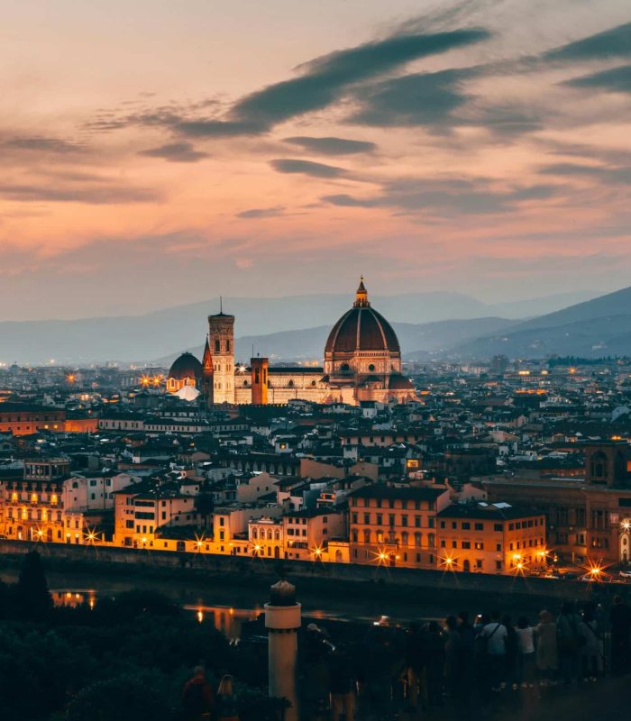 Paysage urbain de Florence au coucher du soleil, avec la cathédrale de Florence illuminée et son dôme emblématique, les bâtiments historiques environnants et les collines lointaines sous un ciel coloré.