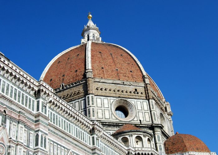 L'image montre le dôme en tuiles rouges de la cathédrale de Florence (Il Duomo) sur fond de ciel bleu clair, avec une façade détaillée en marbre blanc, vert et rose, avec des fenêtres arquées et des motifs complexes.