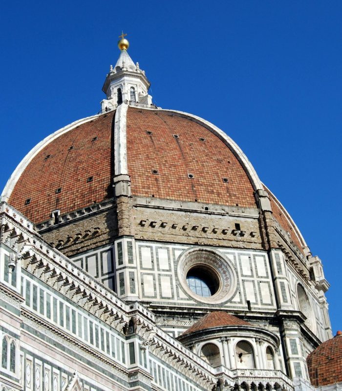 L'image montre le dôme en tuiles rouges de la cathédrale de Florence (Il Duomo) sur fond de ciel bleu clair, avec une façade détaillée en marbre blanc, vert et rose, avec des fenêtres arquées et des motifs complexes.