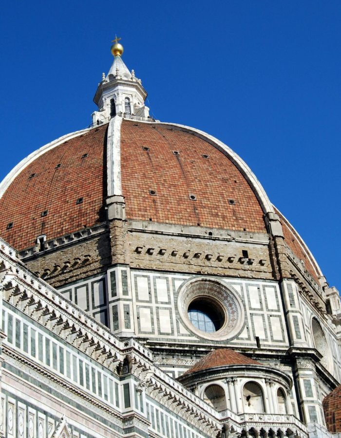 L'image montre le dôme en tuiles rouges de la cathédrale de Florence (Il Duomo) sur fond de ciel bleu clair, avec une façade détaillée en marbre blanc, vert et rose, avec des fenêtres arquées et des motifs complexes.