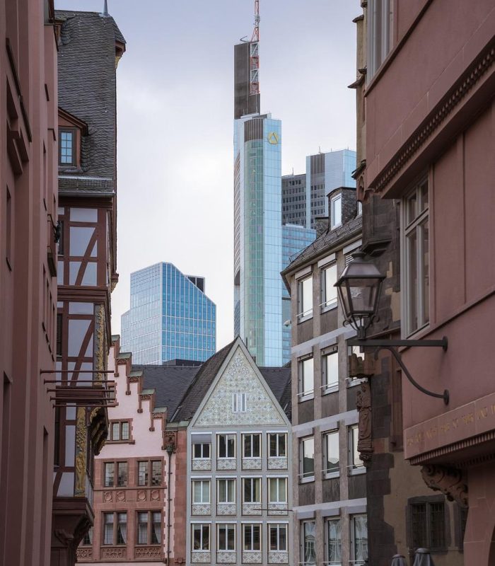 Une rue étroite bordée de bâtiments historiques mène à des gratte-ciel modernes en verre à l'arrière-plan, mêlant architecture ancienne et nouvelle sous un ciel nuageux.