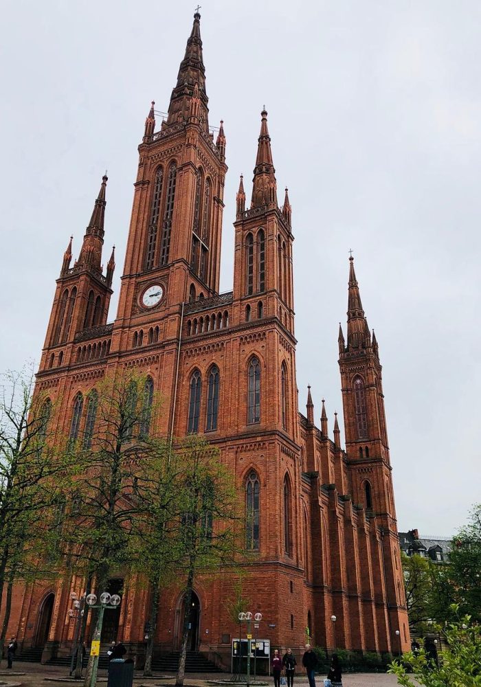 Une grande église de style gothique en briques rouges, dotée de plusieurs flèches et d'une horloge proéminente, fait face à un ciel nuageux ; des arbres verts et des personnes sont visibles à la base de l'édifice.