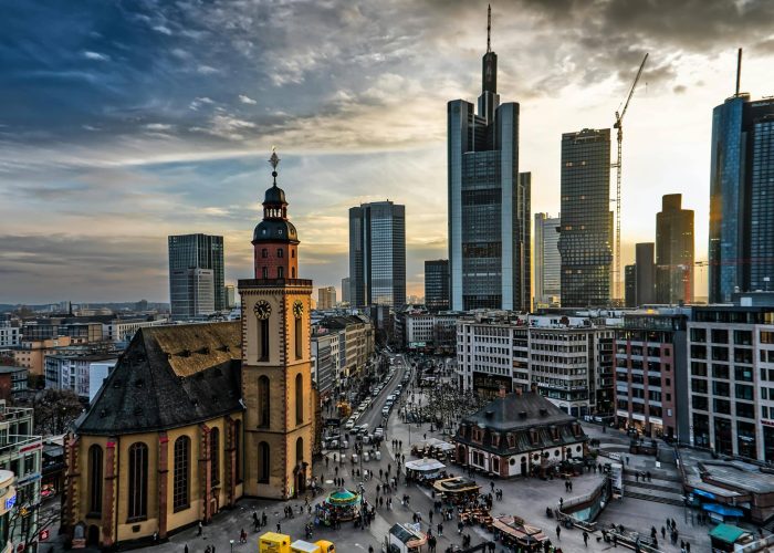 Un paysage urbain au coucher du soleil montre une église historique au premier plan, entourée de foules et de bâtiments, avec des gratte-ciel modernes et des grues de construction à l'arrière-plan.