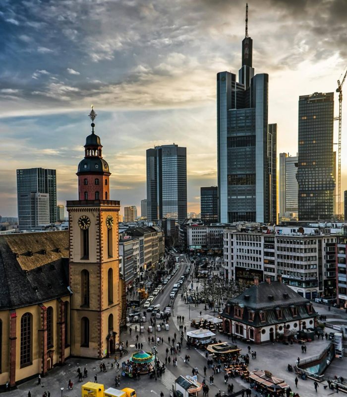 A cityscape featuring a mix of historic and modern architecture, with a church in the foreground, busy streets full of people and tall glass skyscrapers in the background, under a dramatic, cloudy sky at sunset.