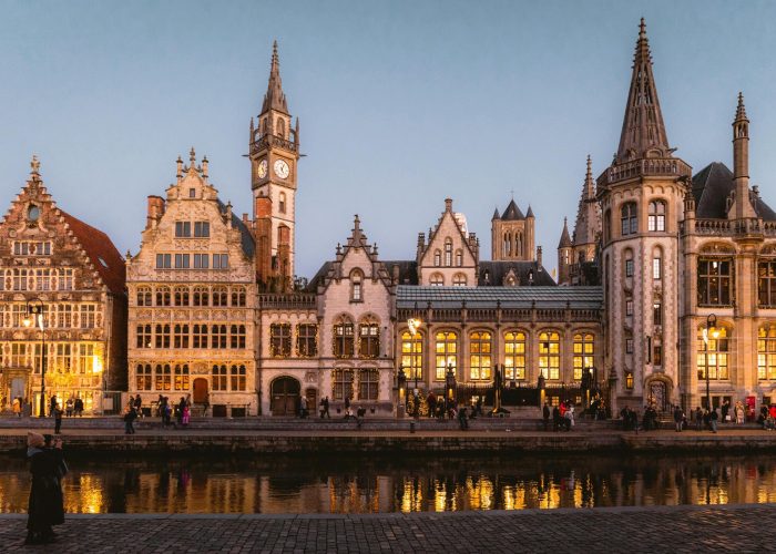 Une rangée de bâtiments historiques ornés de toits pointus et une tour d'horloge sont illuminés au crépuscule le long d'un canal calme à Gand, en Belgique ; les gens se promènent au bord de l'eau.