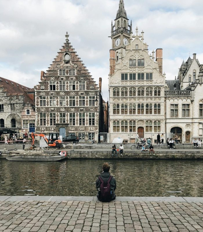 Une personne munie d'un sac à dos est assise au bord d'un canal, face à des bâtiments historiques ornés de pignons en escalier et d'une tour d'horloge dans une ville européenne. Plusieurs personnes marchent et font du vélo le long du front de mer.