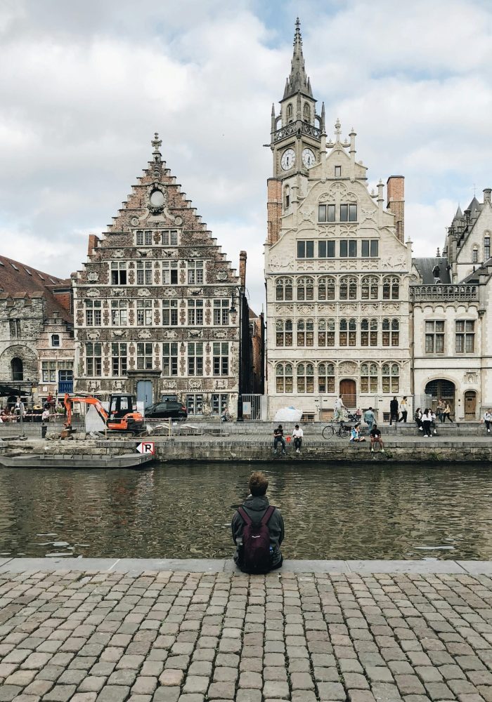 Une personne munie d'un sac à dos est assise au bord d'un canal, face à des bâtiments historiques ornés de pignons en escalier et d'une tour d'horloge dans une ville européenne. Plusieurs personnes marchent et font du vélo le long du front de mer.