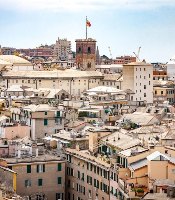Paysage urbain de Gênes, en Italie, avec des bâtiments historiques densément peuplés, aux façades claires et aux toits de tuiles, et une grande tour avec un drapeau italien au centre. Des bâtiments plus modernes et des grues sont visibles à l'arrière-plan.