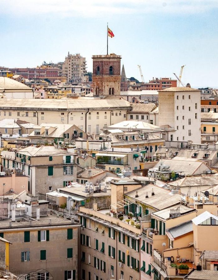 Paysage urbain de Gênes, en Italie, avec des bâtiments historiques densément peuplés, aux façades claires et aux toits de tuiles, et une grande tour avec un drapeau italien au centre. Des bâtiments plus modernes et des grues sont visibles à l'arrière-plan.