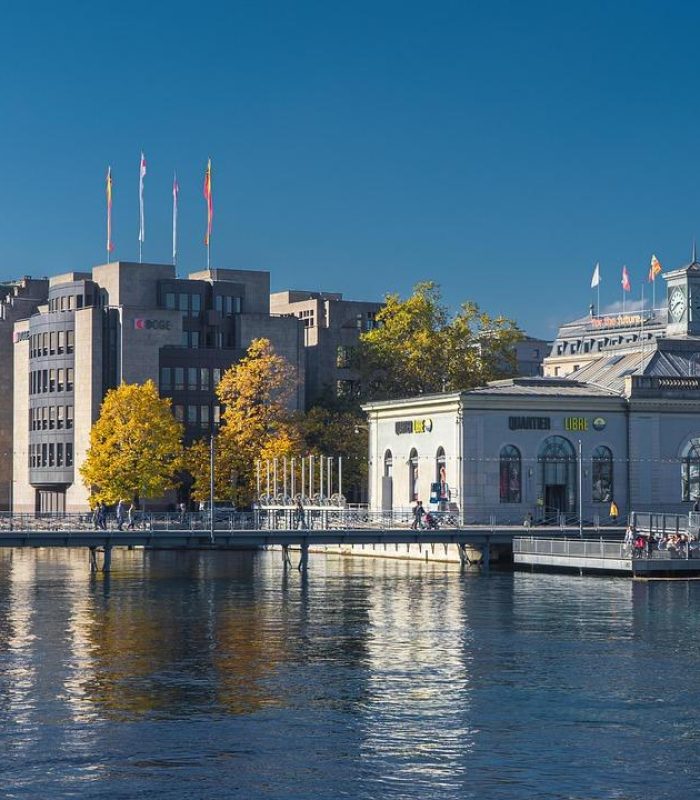 Vue du front de mer sur d'élégants bâtiments historiques et une structure moderne à Genève, en Suisse, sous un ciel bleu clair, avec des arbres et leurs reflets visibles dans l'eau calme.