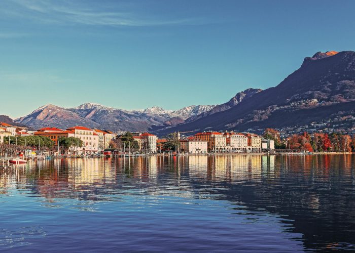 Une paisible ville lacustre aux bâtiments colorés et aux toits rouges se reflète dans une eau calme, avec des rues bordées d'arbres et de hautes montagnes enneigées en arrière-plan, sous un ciel bleu limpide.