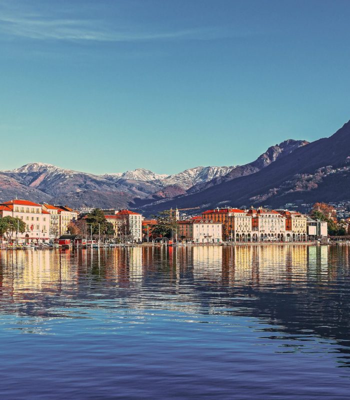 Une paisible ville lacustre aux bâtiments colorés et aux toits rouges se reflète dans une eau calme, avec des rues bordées d'arbres et de hautes montagnes enneigées en arrière-plan, sous un ciel bleu limpide.