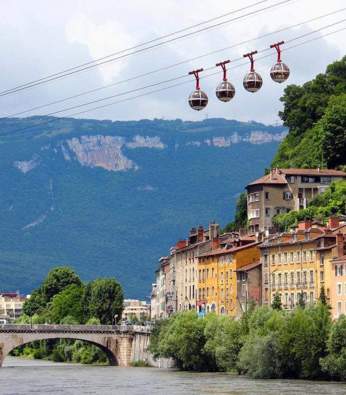A row of colourful buildings flanks a river, crossed by a stone bridge. Four spherical cable cars glide above, surrounded by green hills and steep mountains under a partly cloudy sky.
