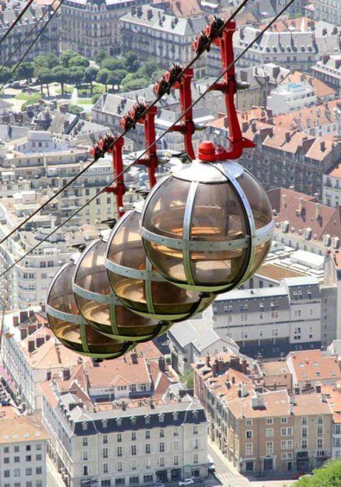 Four spherical glass cable cars circulate above a city of red roofs and tree-lined streets, offering an aerial view of the urban landscape.