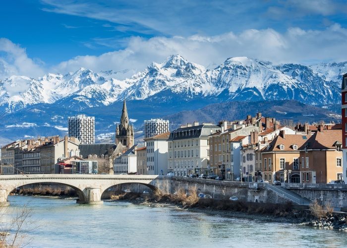A picturesque townscape of colourful buildings, a stone bridge spanning a river and a church with a high spire, set against a backdrop of snow-capped mountains under a blue sky.