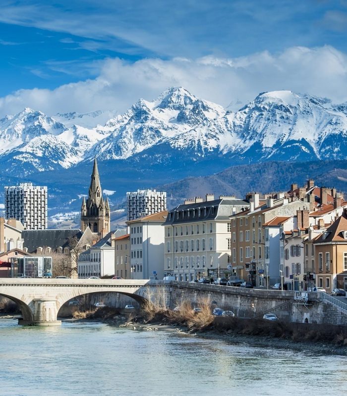 Un paysage urbain pittoresque composé de bâtiments colorés, d'un pont de pierre enjambant une rivière et d'une église avec une haute flèche, sur fond de montagnes enneigées sous un ciel bleu.