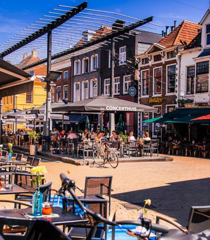 Scène de café en plein air sur une place européenne animée, avec des gens qui mangent et discutent. Des parapluies colorés et des bâtiments historiques bordent la rue sous un ciel ensoleillé. Des tables vides et des vélos sont visibles au premier plan.