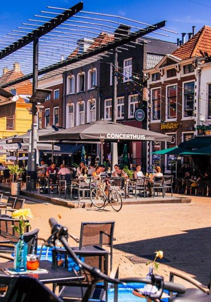 Scène de café en plein air sur une place européenne animée, avec des gens qui mangent et discutent. Des parapluies colorés et des bâtiments historiques bordent la rue sous un ciel ensoleillé. Des tables vides et des vélos sont visibles au premier plan.