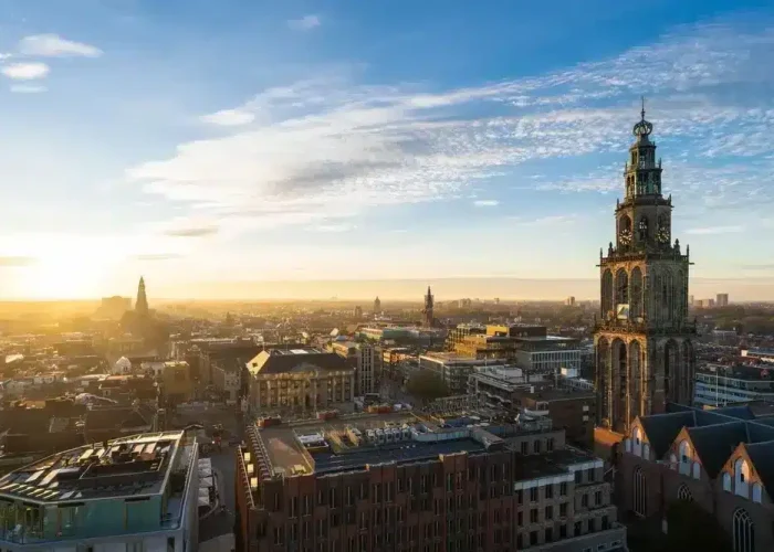 Vue aérienne d'une ville au lever du soleil, avec au premier plan la tour proéminente d'une église historique et un mélange de bâtiments anciens et modernes s'étendant sous un ciel partiellement nuageux.