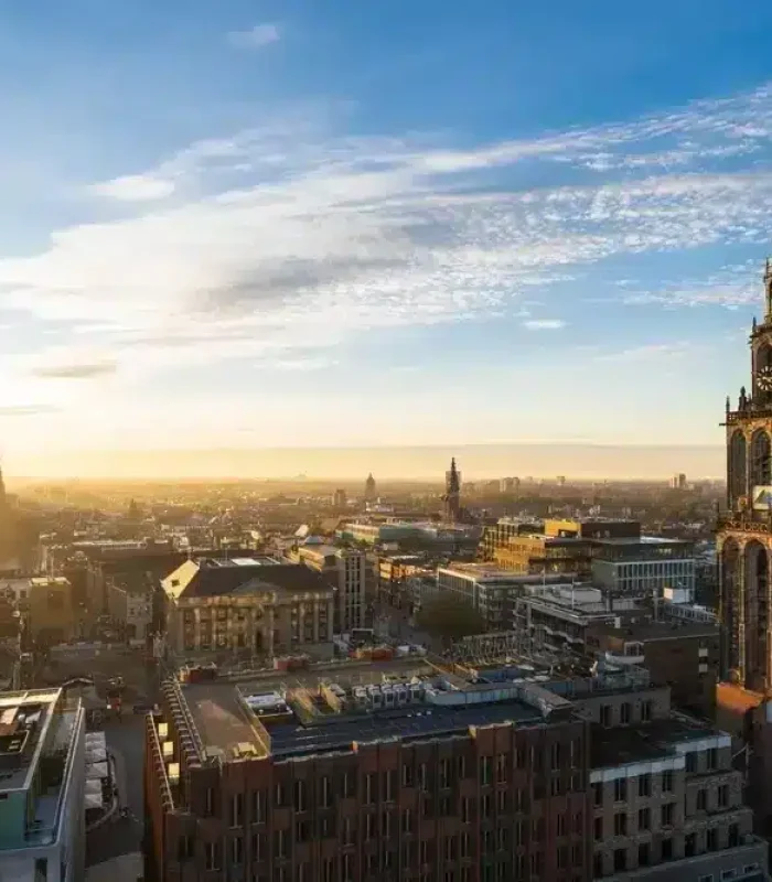 Vue aérienne d'une ville au lever du soleil, avec au premier plan la tour proéminente d'une église historique et un mélange de bâtiments anciens et modernes s'étendant sous un ciel partiellement nuageux.