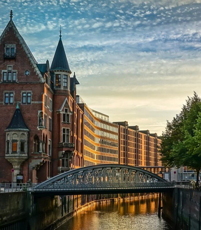 Historic brick building with pointed towers next to a canal and iron bridge, modern buildings in the background, trees on the right, and partly cloudy sky at sunset reflected on the water.