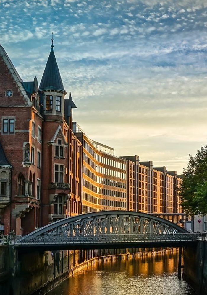 Historic brick building with pointed towers next to a canal and iron bridge, modern buildings in the background, trees on the right, and partly cloudy sky at sunset reflected on the water.
