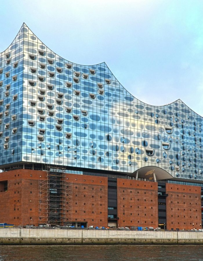 Un bâtiment moderne en verre avec un toit en forme de vague se trouve au sommet d'un vieil entrepôt en briques au bord de l'eau, sous un ciel nuageux. D'autres bâtiments et une tour cylindrique sont visibles à l'arrière-plan.