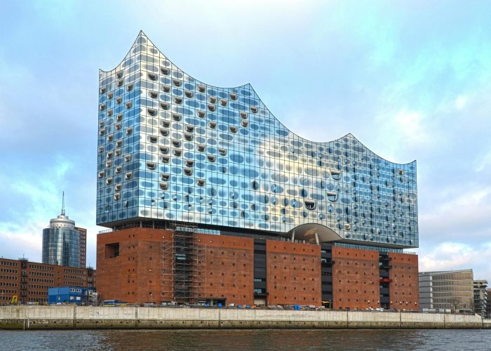A modern glass building with a wave-shaped roof sits atop an old brick warehouse on the water's edge, under a cloudy sky. Other buildings and a cylindrical tower can be seen in the background.