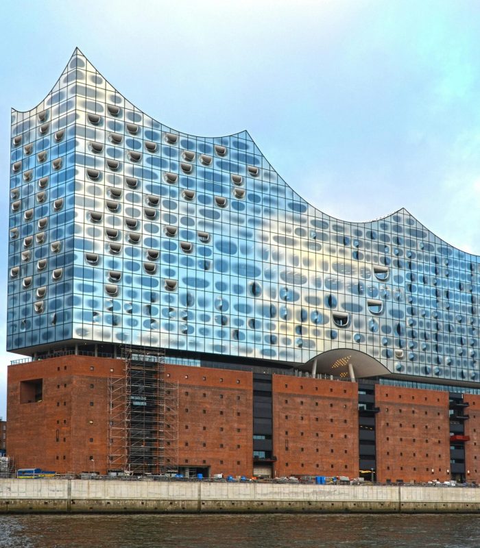Un bâtiment moderne en verre avec un toit en forme de vague se trouve au sommet d'un vieil entrepôt en briques au bord de l'eau, sous un ciel nuageux. D'autres bâtiments et une tour cylindrique sont visibles à l'arrière-plan.