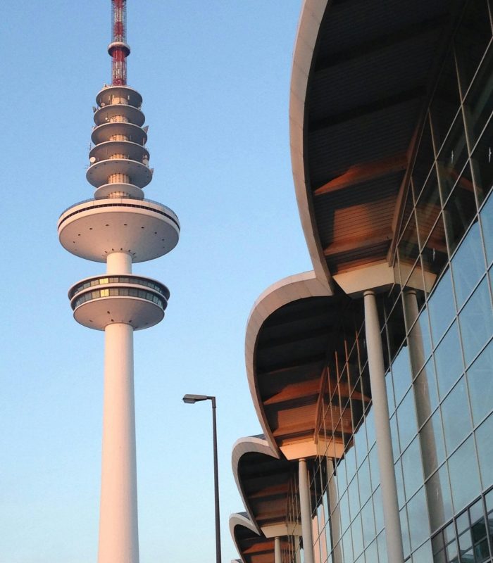 Une tour de télévision avec des ponts d'observation circulaires se détache sur un ciel bleu, à côté d'un bâtiment moderne avec un toit incurvé et une façade en verre reflétant la lumière du soleil. Un lampadaire est visible au premier plan.