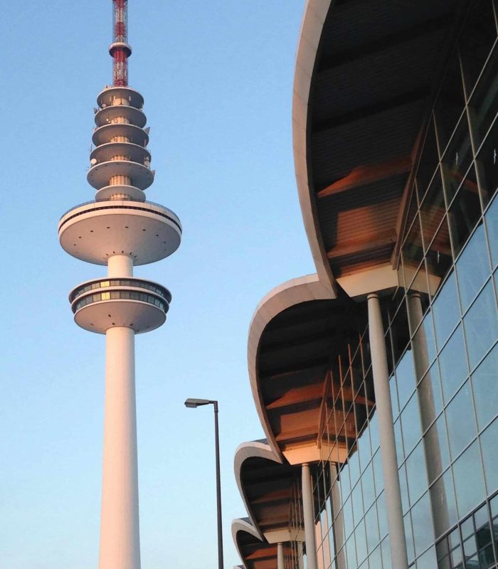 Une tour de télévision avec des ponts d'observation circulaires se détache sur un ciel bleu, à côté d'un bâtiment moderne avec un toit incurvé et une façade en verre reflétant la lumière du soleil. Un lampadaire est visible au premier plan.