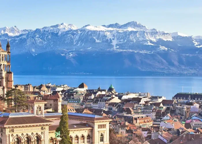 Vue panoramique de Lausanne, en Suisse, montrant des bâtiments historiques, une cathédrale, le lac Léman et les Alpes enneigées en arrière-plan, sous un ciel bleu clair.