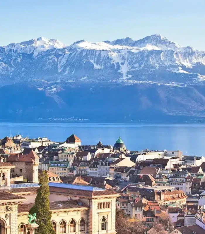 Vue panoramique de Lausanne, en Suisse, montrant des bâtiments historiques, une cathédrale, le lac Léman et les Alpes enneigées en arrière-plan, sous un ciel bleu clair.